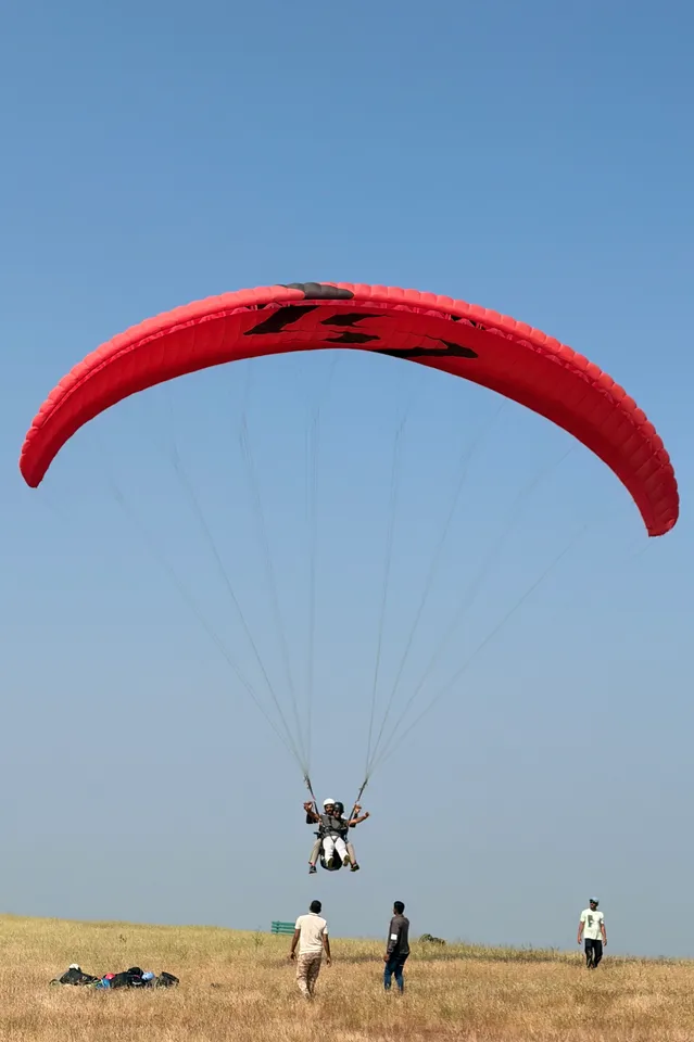A red paraglider lifting above the take-off field with people below