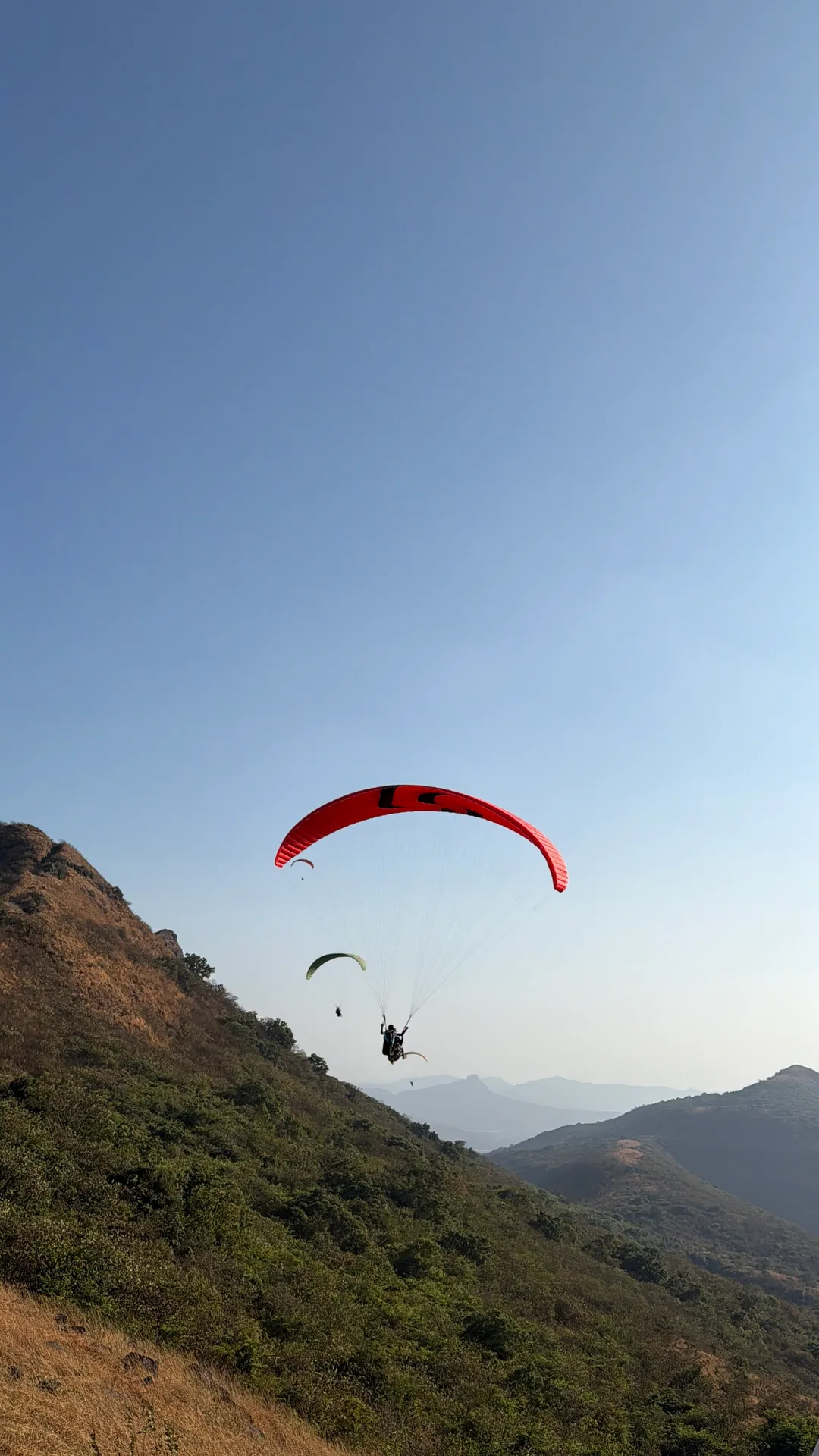 A red paraglider flying beside a green hillside above the valley