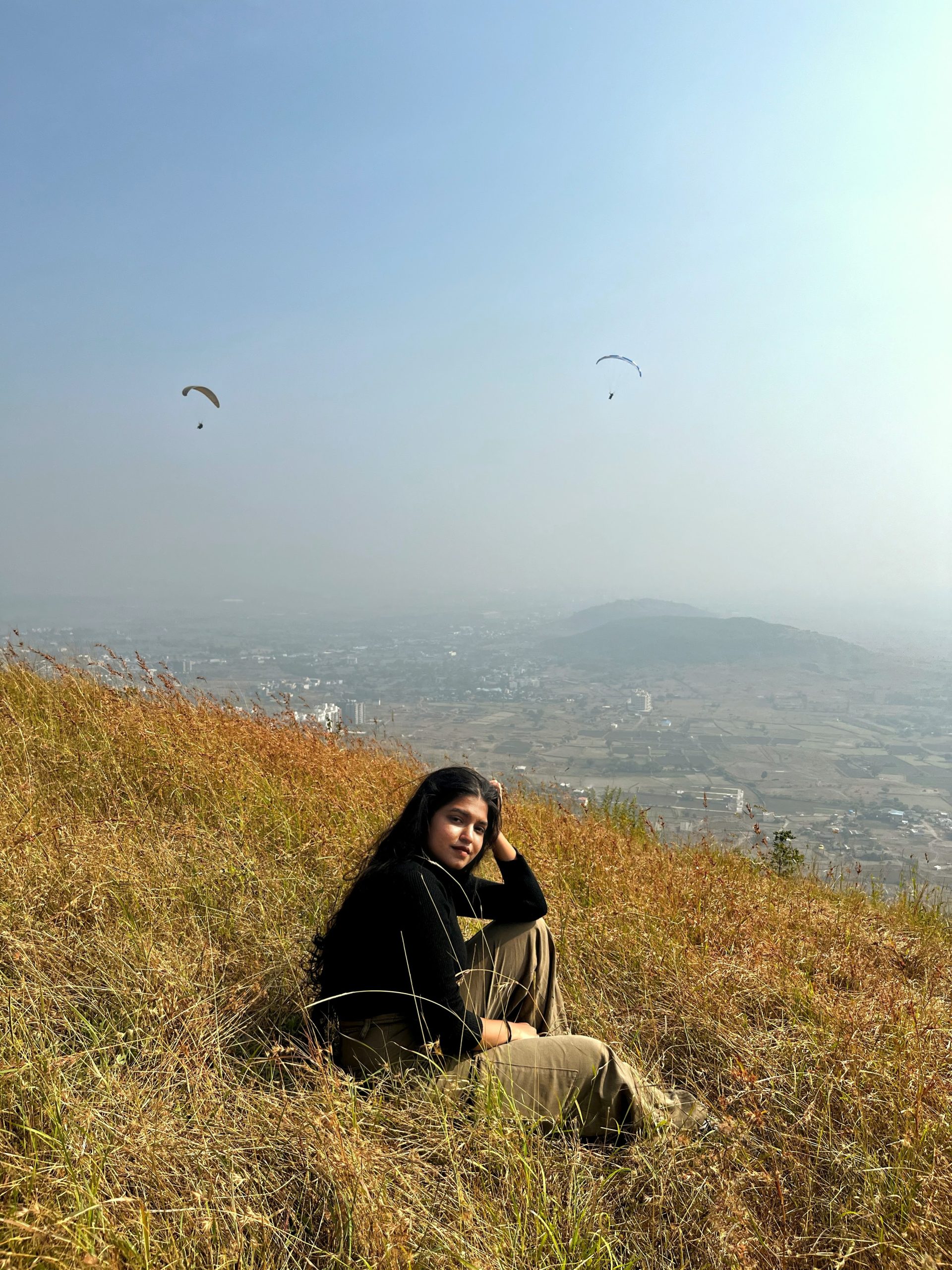 a woman sitting on a hill with a view of a city and paragliders